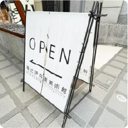 白地に黒文字を配置した黒い細身フレームの美術館のスタンド看板