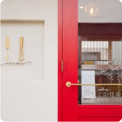 白壁と赤い扉にマッチした、ゴールド調のミニマルな文字看板