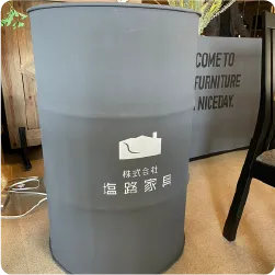 グレーに着色したドラム缶に白色の会社名とロゴを配したサイン